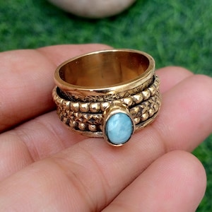 Pode incluir: Um anel giratório largo de latão com uma pedra oval azul-clara no centro. A banda é composta por várias camadas texturizadas, incluindo uma borda com contas e um padrão gravado decorativo, criando uma aparência metálica em camadas.