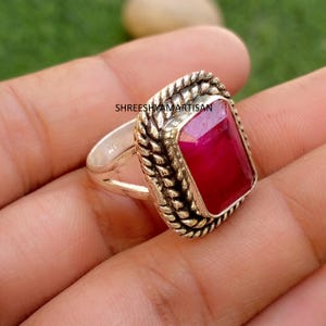 May include: A silver ring featuring a rectangular, deep red gemstone set in a frame with a twisted rope texture. The band is split into two thin silver segments. The text SHREESHYAMARTISAN is visible on the metal surface near the gemstone.