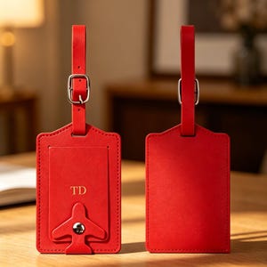 May include: Two rectangular red luggage tags with matching straps and silver-toned metal buckles. One tag features a flap with the letters TD embossed in gold and a small airplane-shaped metal snap closure. The other tag is a solid red rectangle.
