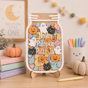 Op de afbeelding: Een decoratief houten bord in de vorm van een pot met de tekst Noah's Pumpkin Patch Jar. De binnenkant is gevuld met kleurrijke houten uitsparingen van oranje pompoenen, witte spoken, zwarte katten, gele manen en pastelkleurige bladeren.