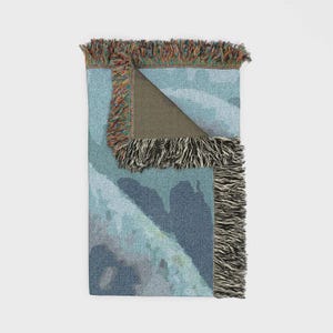 May include: A woven throw blanket featuring a blue and grey abstract landscape pattern. The blanket is folded at the top corner to reveal a solid dark grey underside. The edges are finished with a multi-coloured fringe made of various yarn colours.