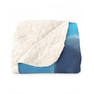 May include: A folded throw blanket featuring a plush, cream-coloured sherpa fleece lining on one side and a smooth, printed exterior with a gradient of blue tones ranging from light sky blue to a deep, dark navy blue across the fabric surface.