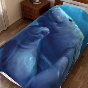 May include: A soft bed blanket featuring a large, detailed print of two manatees swimming together in deep blue water. The artwork displays the grey, rounded bodies of the marine mammals against a gradient of dark and light blue aquatic tones.
