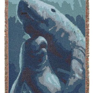 May include: A woven throw blanket featuring a stylised graphic of a shark swimming in deep blue and light blue water. The design uses a pixelated, colour-blocked aesthetic with shades of navy, teal, and white. The blanket has a fringed edge.