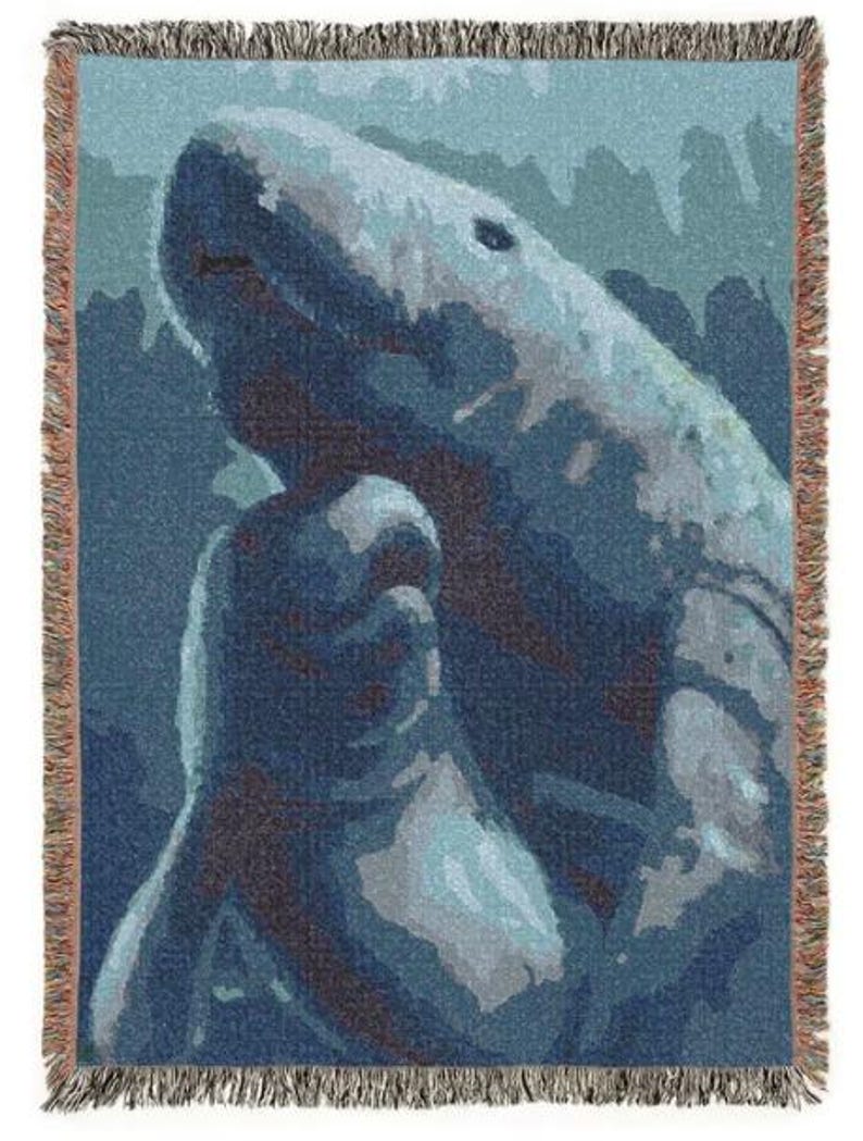 May include: A woven throw blanket featuring a stylised graphic of a shark swimming in deep blue and light blue water. The design uses a pixelated, colour-blocked aesthetic with shades of navy, teal, and white. The blanket has a fringed edge.