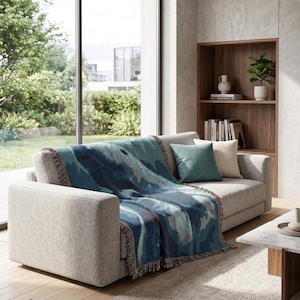 May include: A woven throw blanket featuring an abstract pattern in shades of deep blue, teal, and light grey with fringed edges, draped over a light grey sofa. The blanket displays a fluid, layered design with varied blue and grey colour blocks.