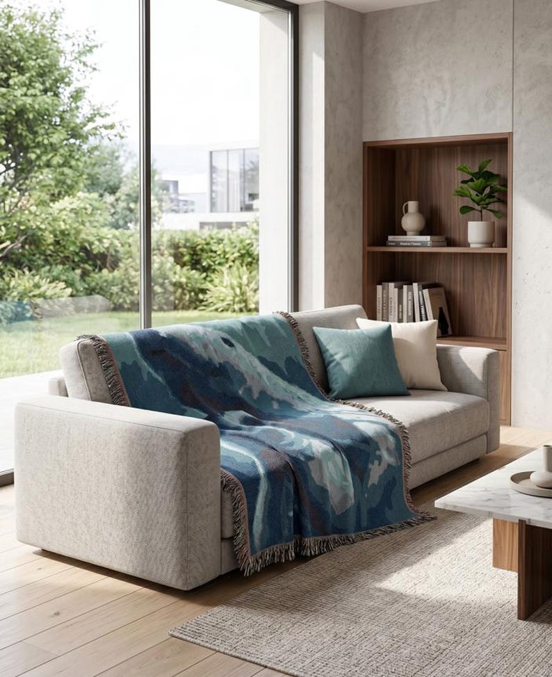 May include: A woven throw blanket featuring an abstract pattern in shades of deep blue, teal, and light grey with fringed edges, draped over a light grey sofa. The blanket displays a fluid, layered design with varied blue and grey colour blocks.