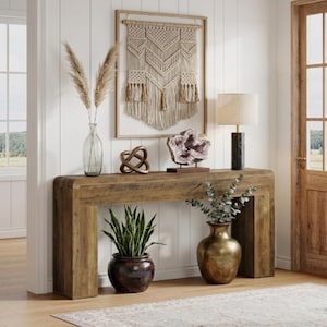 May include: A wooden console table with a smooth, curved frame. On top sits a glass vase with dried grass, a bronze-colored knot sculpture, a large amethyst geode, and a lamp with a black marble base. Below, a snake plant and a gold-toned vase.