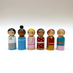 May include: Seven wooden peg dolls, each with a different painted design and hairstyle. The dolls are standing in a row on a white surface.