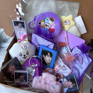 May include: A cardboard gift box filled with purple and pink themed merchandise, including a plush coin purse with cartoon characters, a star-shaped plush, a blue-framed photo, a fuzzy pink accessory, and several small cards featuring a person's portrait.