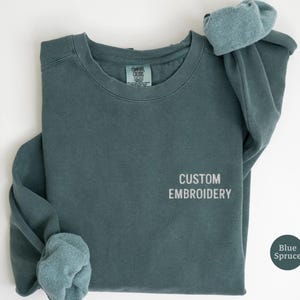 May include: A blue spruce colored crewneck sweatshirt featuring the text CUSTOM EMBROIDERY stitched in a light gray thread on the right chest area. The garment has long sleeves with ribbed cuffs and a Comfort Colors brand label at the neckline.