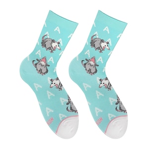 Könnte beinhalten: Ein Paar hellblaue Socken mit einem sich wiederholenden Pixel-Art-Muster aus grauen und weißen Opossums mit rosa Akzenten und dem Buchstaben A. Die Socken haben einen einfarbig weißen Zehenbereich und ein geripptes Bündchen am oberen Rand.
