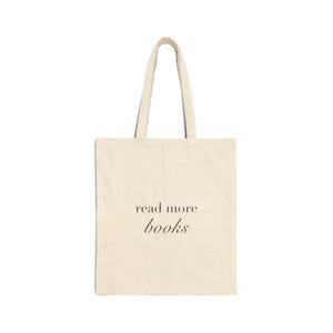 Könnte beinhalten: Eine cremefarbene Stofftasche aus Canvas mit zwei Schulterriemen. Auf der Vorderseite ist der Text "read more books" in einer dunklen Serifenschrift aufgedruckt, wobei "read more" in der oberen Zeile und "books" kursiv darunter steht.