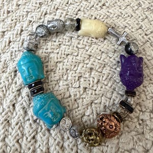 May include: A stretch bracelet featuring a variety of beads including two turquoise-colored Buddha heads, a purple Buddha head, a cream-colored Buddha head, a silver-toned cross, metallic lion heads, and small black and silver accent beads.