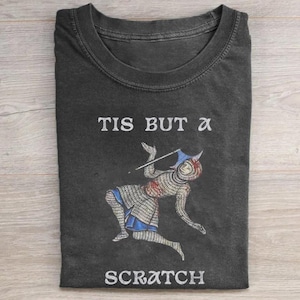 May include: A charcoal gray t-shirt featuring a graphic of a medieval knight in chainmail armor with a blue helmet and a spear. The text "TIS BUT A SCRATCH" is printed in white capital letters above and below the knight illustration.