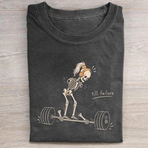 May include: A charcoal gray t-shirt featuring a graphic of a skeleton wearing orange headphones and a ponytail, lifting a barbell. The text "till failure" is printed in white script next to the skeleton on the front of the garment.