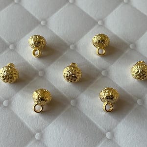 以下が含まれることがあります： 6つの小さな丸い金色の金属製チャーム。それぞれに質感のあるエンボス加工が施されており、ベースには取り付け用の円形のループが1つ付いています。チャームは白い背景に対して散らばるように配置されています。