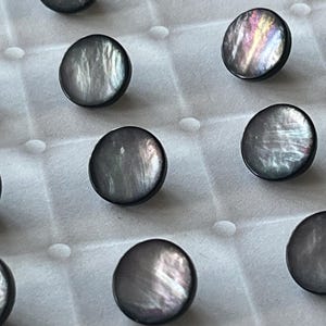 Puede incluir: Una colección de botones redondos con una superficie oscura e iridiscente con tonos grises y morados. Cada botón tiene un acabado liso y pulido con un brillo distintivo y resplandeciente que refleja la luz sobre la superficie circular.