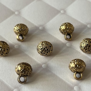 以下が含まれることがあります： 6つの小さな丸い金色の金属製ボタンのセット。各ボタンの表面には、詳細な隆起した渦巻き状のフィリグリー模様が施されています。3つのボタンには、縫い付け用の小さな円形のループが底面に付いています。