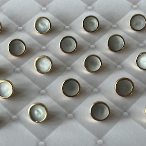 May include: A collection of seventeen circular buttons arranged in a grid pattern. Each button features a smooth, pale grey centre with a subtle pearlescent sheen, encased within a thin, polished gold-toned metal rim that creates a distinct border.