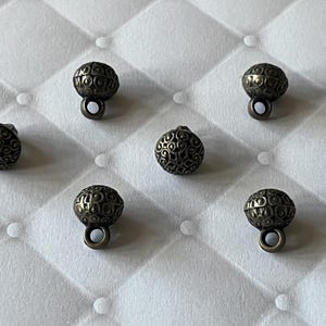 May include: A collection of seven small, spherical metal buttons with an antique bronze finish. Each button features a detailed, raised filigree pattern across its surface and includes a small, circular loop at the base for attachment.