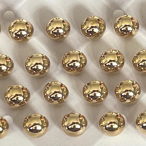 May include: A collection of twenty-three small, spherical, metallic gold beads arranged in a grid pattern. Each bead features a smooth, highly reflective surface that catches the light, creating bright highlights across the rounded, polished gold exterior.