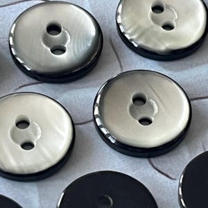 Op de afbeelding: Een verzameling ronde knopen met een glanzend, gebroken wit midden met een subtiele parelmoerglans, omringd door een opvallende zwarte buitenrand. Elke knoop is ontworpen met twee centrale gaten voor bevestiging en heeft een glad oppervlak.