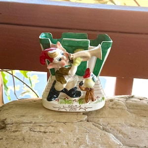 Op de afbeelding: Een grillige keramische plantenbak met een cartoon-elf die op een hoorn blaast, met een vogel figuur ernaast. De plantenbak heeft een groen en wit kasteelachtig ontwerp. De elf draagt een rode hoed, groene handschoenen en zwarte laarzen. De basis is wit met bloemaccenten.