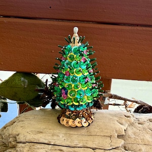 May include: A decorative cone-shaped ornament resembling a tree, covered in shimmering green and pink sequins with protruding dark green pins. The base is adorned with metallic bronze sequins, and a single white bead sits at the very top point.