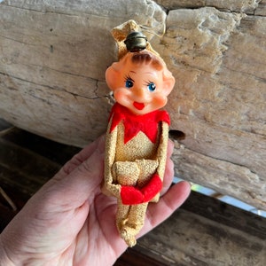 May include: A vintage elf ornament featuring a moulded plastic face with blue eyes and red lips. The elf wears a gold textured fabric suit with a red collar and red mittens. A small metal bell is attached to the top of the gold fabric hat.