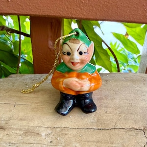 Könnte beinhalten: Ein Keramik-Elfenornament mit einer grünen spitzen Mütze, einem orangefarbenen Hemd und schwarzen Stiefeln. Der Elf hat ein bemaltes Gesicht mit geschwungenen Augenbrauen, rosigen Wangen und einem Lächeln. Eine goldene Schnur ist an der Mütze befestigt.
