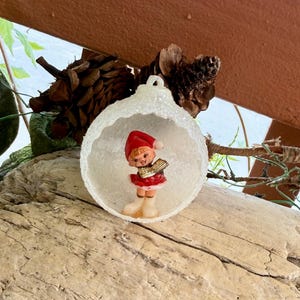 May include: A Christmas ornament featuring a small figurine of a girl in a red Santa hat and dress, holding a book. The figurine is inside a white, glittery, open sphere. The ornament is set on a piece of wood.