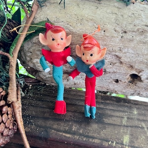 May include: Two vintage-style elf ornaments with moulded plastic heads, pointed ears, and painted facial features. One elf wears a red collar and teal felt body, while the other wears a teal collar and red felt body. Both have small hanging loops.
