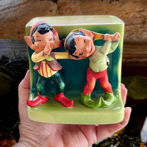 Puede incluir: Una jardinera de cerámica vintage con dos figuras de duendes. La jardinera tiene forma rectangular con una base verde y un fondo verde oscuro. Los duendes visten trajes coloridos con detalles rojos, verdes y amarillos.