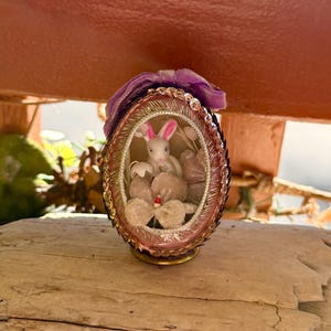 以下が含まれることがあります： ピンクとゴールドのフレームのイースターエッグオーナメント。ピンクの耳を持つ白いウサギが中にいます。卵はスパンコールと紫のリボンで飾られています。オーナメントは木製の表面に置かれています。