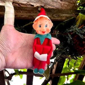 May include: A vintage Christmas elf figurine. The elf has a red felt outfit, a green collar, and a white-trimmed red hat. The elf's face is made of plastic with blue eyes and a cheerful expression. The elf is holding a white book.