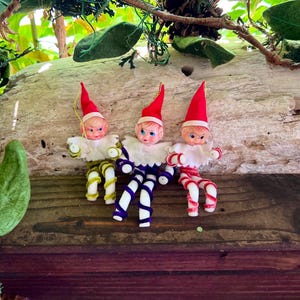 May include: Three small elf ornaments with pointed red hats and white collars. Each elf has a plastic face with painted features and limbs made of coiled pipe cleaners in olive green, dark purple, and red and white stripes respectively.
