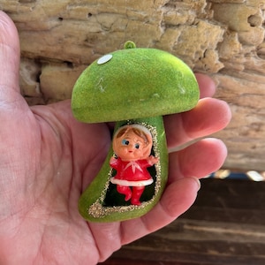 Puede incluir: Adorno de hongo vintage con una tapa y tallo verdes aterciopelados. Una pequeña muñeca con un atuendo rojo está dentro del tallo. El adorno tiene un borde dorado brillante y un punto blanco en la tapa.