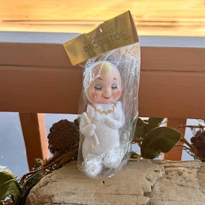 Puede incluir: Una figura de muñeco de estilo vintage con un conjunto de punto blanco y un cuello de color dorado, dentro de una bolsa de plástico transparente con una tarjeta dorada en la parte superior que dice CHRISTMAS DECORATION. Tiene cara pálida.