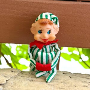 May include: A small elf figurine with a molded plastic face, blue eyes, and a smiling expression. The elf wears a green and white striped fabric hat and matching striped pajamas with a red collar and red cuffs on the sleeves and feet.