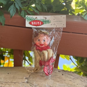 May include: A Shiny Brite Christmas Pixie ornament in original plastic packaging. The pixie has a painted face, red lips, and a gold fabric body with red accents. The header card features the text Shiny Brite Christmas Pixie and Japan.