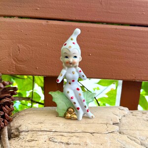 May include: A ceramic figurine of a seated elf wearing a white suit with red and gold polka dots. The elf has a pointed hat and is positioned on a green holly leaf base with three gold-colored berries at the bottom. The figure has painted facial features.