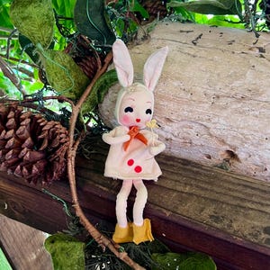 以下が含まれることがあります： ピンクの耳と黄色の靴を履いた白いウサギのぬいぐるみが木製の表面に座っています。ぬいぐるみは赤いボタンと首に赤いリボンが付いたピンクのドレスを着ています。ぬいぐるみは手に黄色の花を持っています。