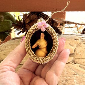 以下が含まれることがあります： 金色のイースターエッグのオーナメント。中には小さな黄色のウサギの置物が入っています。卵は金のチェーンで縁取られ、ピンクの花で飾られています。吊り下げ用の金の紐が付いています。