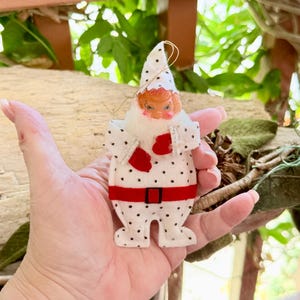 Op de afbeelding: Een witte kerstversiering van vilt in de vorm van een figuur met een puntmuts, voorzien van kleine zwarte stippen, een rode riem met een zwarte gesp en rode wanten. Het figuur heeft een gevormd plastic gezicht met een witte baard.