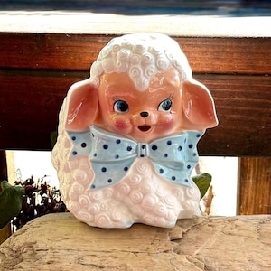 Könnte beinhalten: Eine Lammfigur aus Keramik mit weißer, lockiger Wollstruktur, rosa Ohren und Wangen sowie leuchtend blauen Augen. Das Lamm trägt eine große, hellblaue Schleife mit dunkelblauen Punkten um den Hals, was für ein dekoratives und detailliertes Aussehen sorgt.