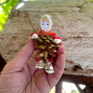 May include: A vintage Christmas ornament in the shape of a skier. The skier's body is a gold-painted pinecone, with a doll head wearing a white knit cap and orange scarf. Red ski poles and white pom-pom mittens complete the look.