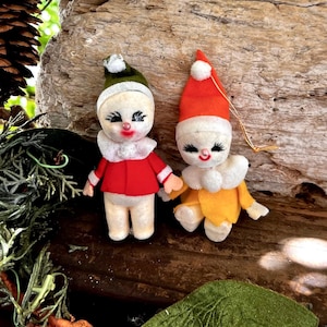 May include: Two vintage Christmas elf ornaments. One elf wears a red jacket and green hat, the other wears a yellow jacket and red hat. Both have white collars, rosy cheeks, and black eyelashes. They are made of felt and have a whimsical design.
