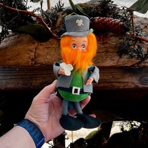 May include: A vintage St. Patrick's Day leprechaun doll. The doll has a gray hat with a shamrock, an orange beard, and a green outfit with a black belt and shoes. The doll is holding a white shamrock.