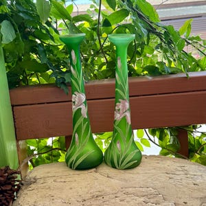 May include: Two tall, slender, frosted green glass vases featuring a painted design of light pink iris flowers with long green stems and leaves. Each vase has a flared, circular rim at the top and a wider, rounded base, standing upright.